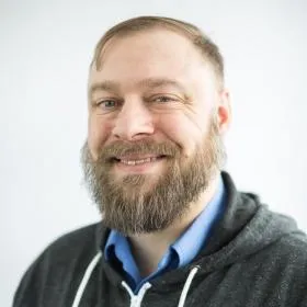 Smiling man with a full beard wearing a hoodie against a light background.