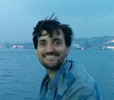 Smiling man with dark hair sitting near the water at dusk with city lights in the background.