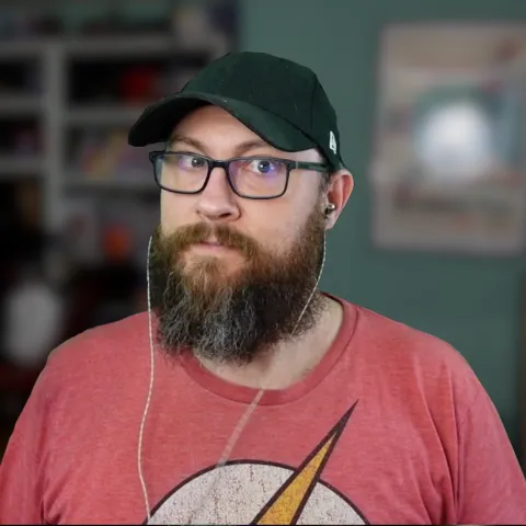 Man with glasses, a beard, and a black cap wearing a Flash logo shirt and earbuds indoors.