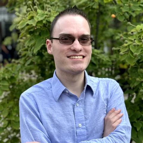 Smiling man with glasses wearing a light blue shirt standing outdoors in front of greenery.
