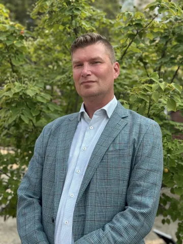 Man in a patterned blazer and light blue shirt standing outdoors in front of leafy trees.