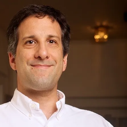 Smiling man wearing a white button-up shirt indoors with a softly blurred background.