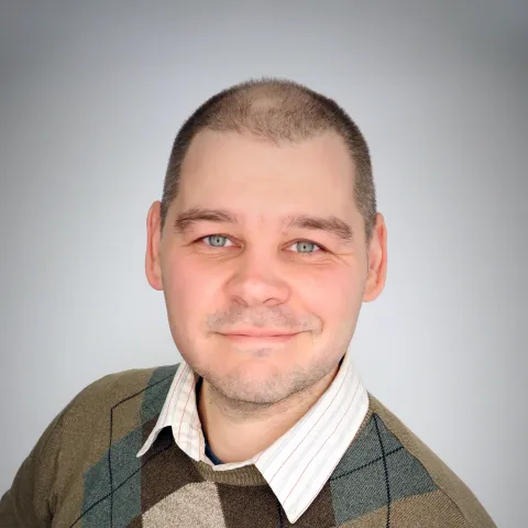 Smiling man with closely cropped hair wearing a collared shirt and patterned sweater against a light background.