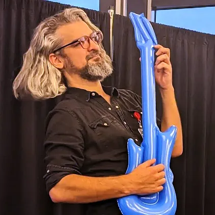 Person with long gray hair and glasses playfully holding a blue inflatable guitar.