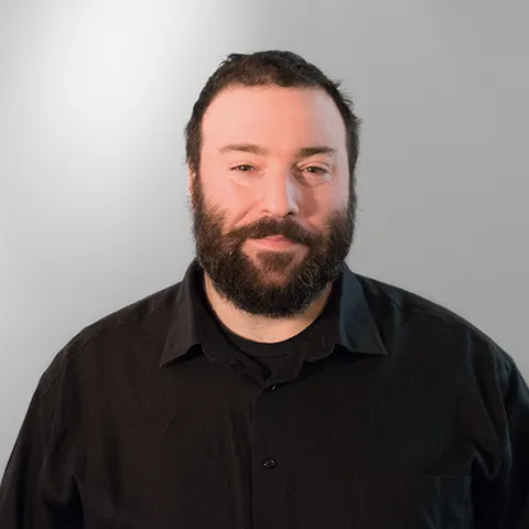 Man with a full beard wearing a black button-up shirt against a light background.