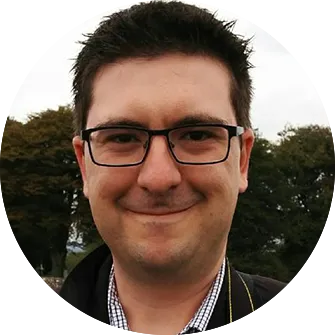 Smiling man with glasses outdoors in front of trees.