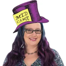 Smiling person with purple and blue hair wearing glasses and a whimsical purple top hat labeled “MidCamp.”