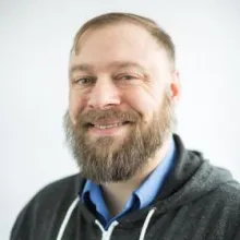 Smiling man with a full beard wearing a hoodie against a light background.