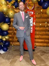 Man in a gray suit with a pink tie and loafers standing in front of festive balloons and a vintage gas pump backdrop.