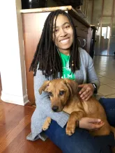 Smiling person with long dreadlocks sitting indoors while holding a brown puppy.