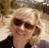 Smiling person with blonde hair and sunglasses outdoors with mountains and trees in the background.
