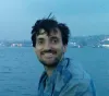 Smiling man with dark hair sitting near the water at dusk with city lights in the background.
