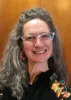 Smiling person with long wavy gray hair and glasses wearing colorful earrings and a floral-patterned top.