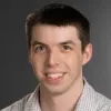 Smiling man with short dark hair wearing a light checkered shirt against a gray background.